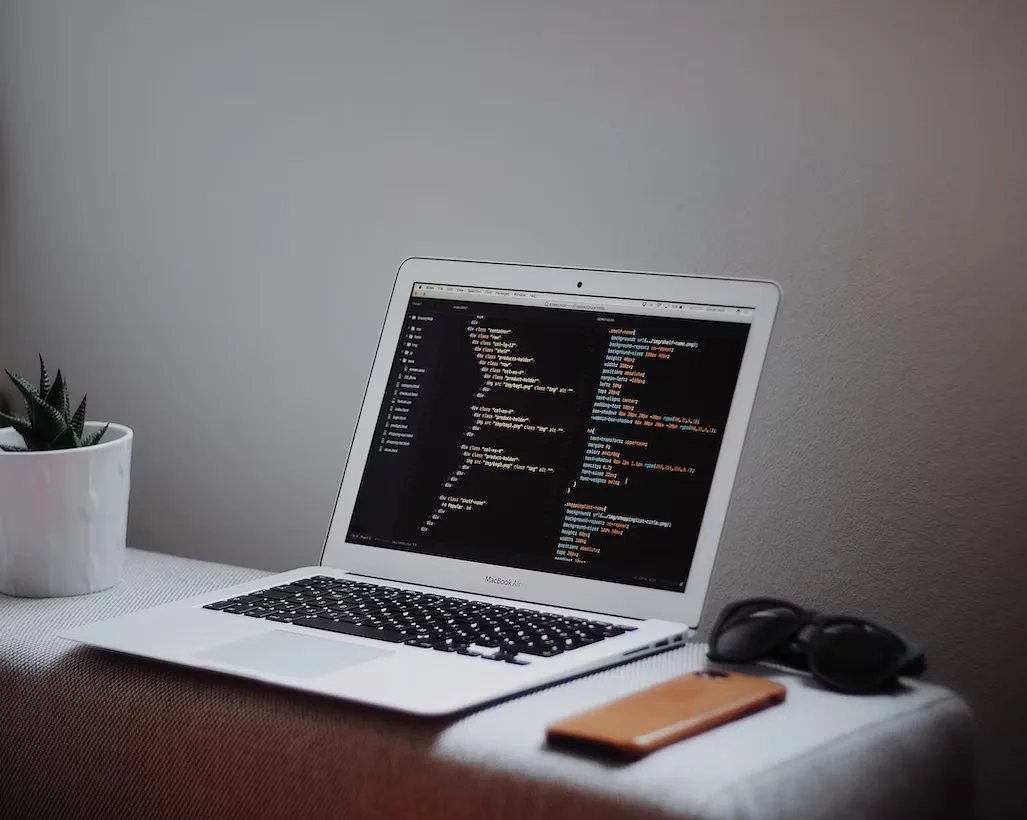 Laptop showing code on a desk beside a phone and a plant